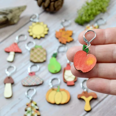 Apple, a handpainted wooden stitch marker, held on hand with autumnal stitch markers in background Apple, a handpainted wooden stitch marker, held on hand with autumnal stitch markers in background